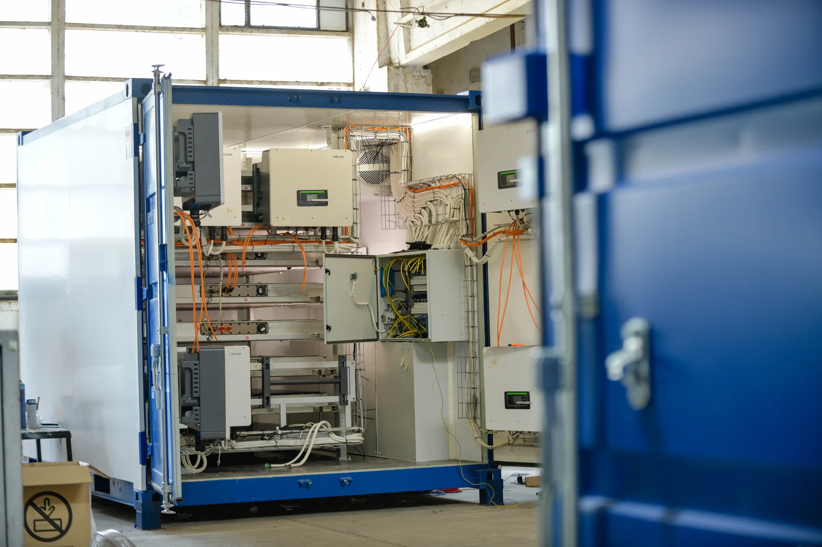 Un Battery-Shelter Marny Energy dans l’atelier, portes ouvertes : conteneur bleu de 10 pieds, onduleurs hybrides, racks de batteries et câblage haute tension